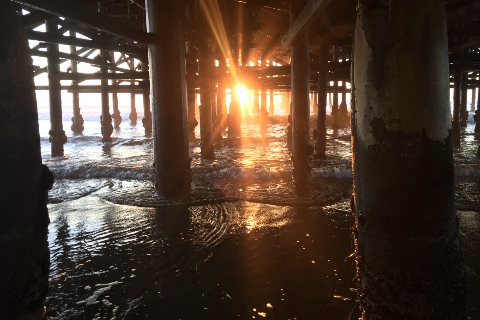 The sun aligned directly beneath Crystal Pier, centred between two receding rows of pilings, reflected on wet sand.